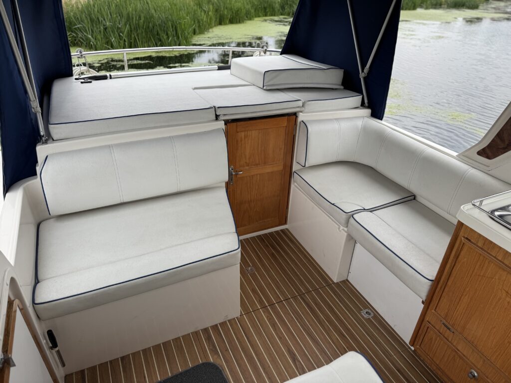 A yacht cockpit with white cushioned bench seating and a teak deck, shown beside calm water and reeds.