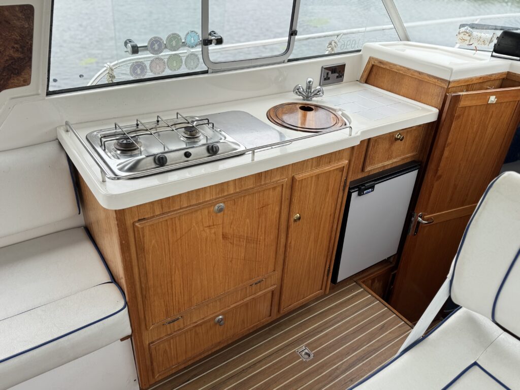 A compact boat galley with a stainless steel two-burner stove, round sink, wood cabinets, and a small refrigerator beside a striped floor mat.