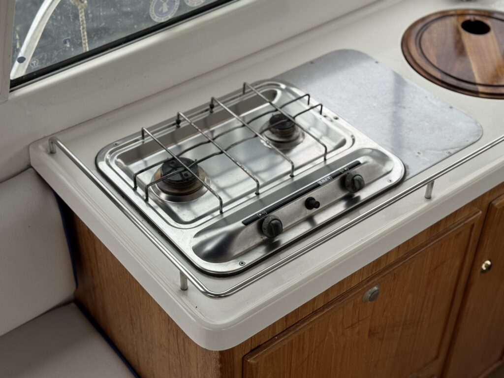 A stainless steel two-burner gas cooktop is built into a white countertop inside a boat galley, with a round wooden sink cover beside it and wood cabinets below.