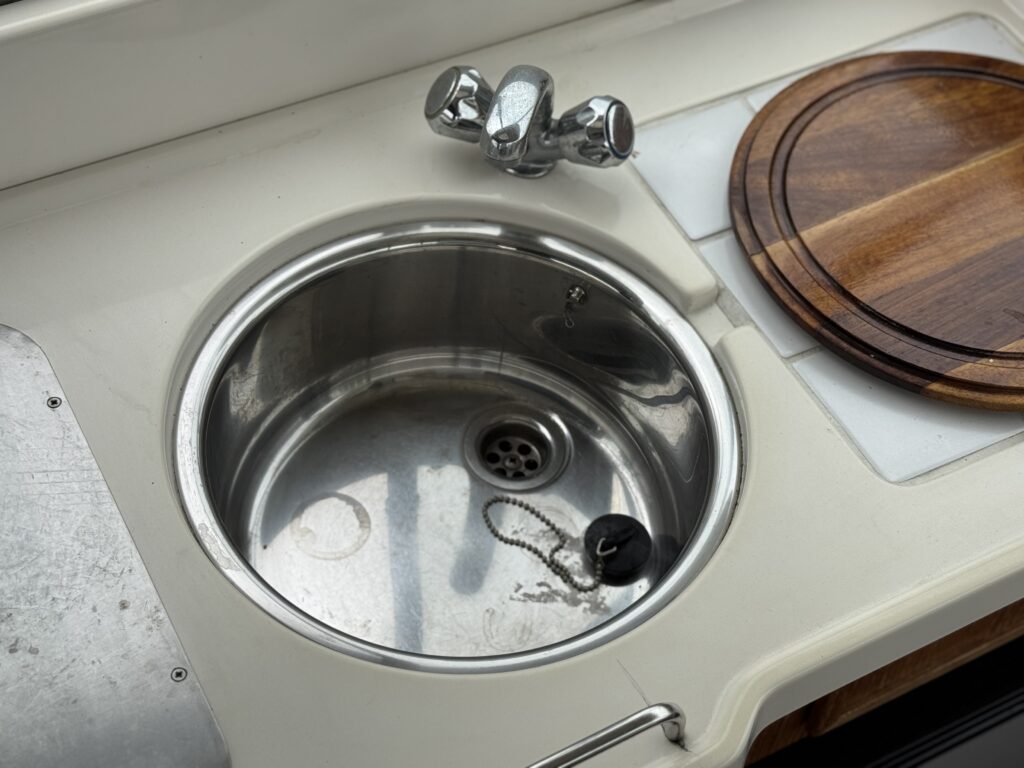 A stainless steel sink with a chrome faucet and drain stopper is set into a white countertop beside a round wooden cutting board.