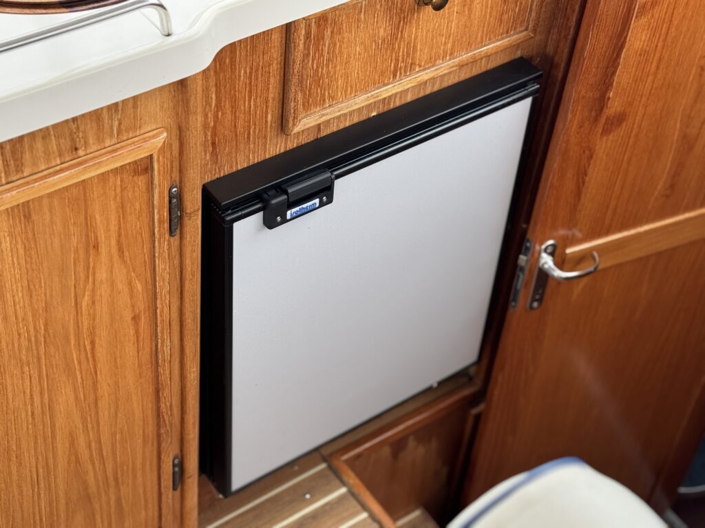 A compact built-in refrigerator with a silver door is installed between wooden cabinets in a boat galley.