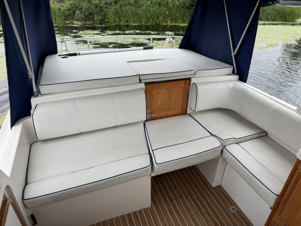 A boat’s covered rear seating area with white cushioned benches around a central hatch, a teak floor, and dark blue canvas sides overlooking the water.