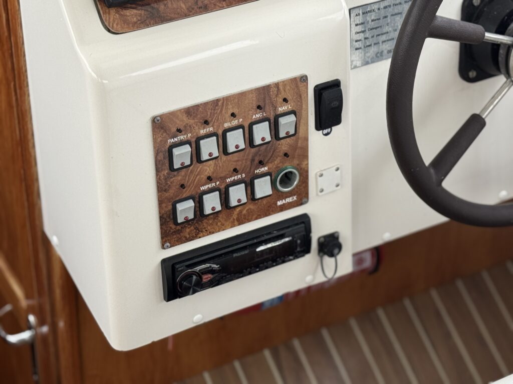 A boat helm console with a wood-grain switch panel, radio unit, and steering wheel, set above a striped floor.