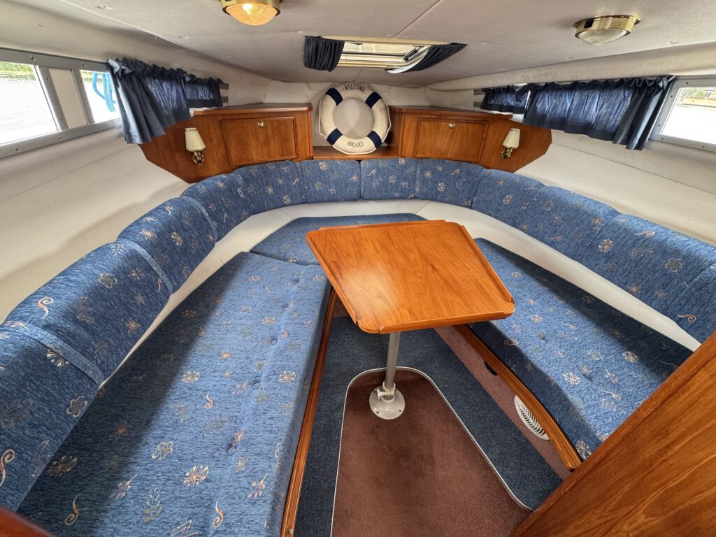A compact boat cabin with blue U-shaped bench seating around a wooden table, framed by wood cabinetry, curtained windows, and a life ring on the rear wall.