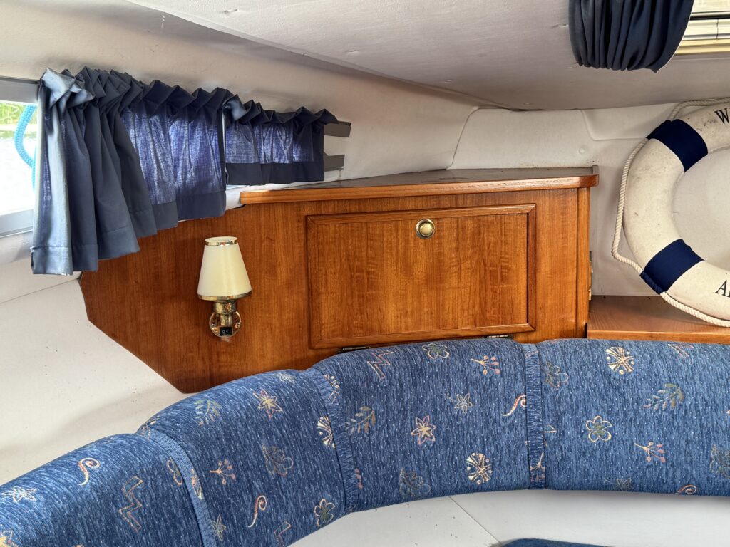 A blue upholstered curved bench in a boat cabin, with a wooden cabinet, small wall lamp, and blue curtains by the window.