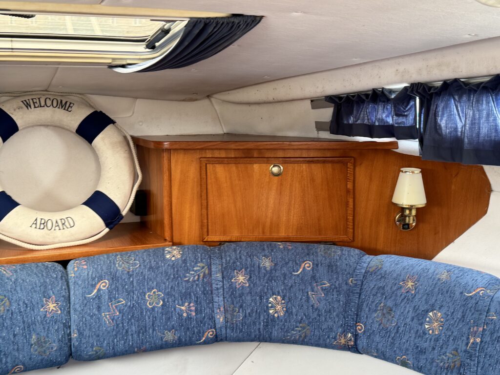 A blue upholstered bench seat inside a boat cabin with a wooden cabinet, a wall lamp, and a life preserver labeled "Welcome Aboard."