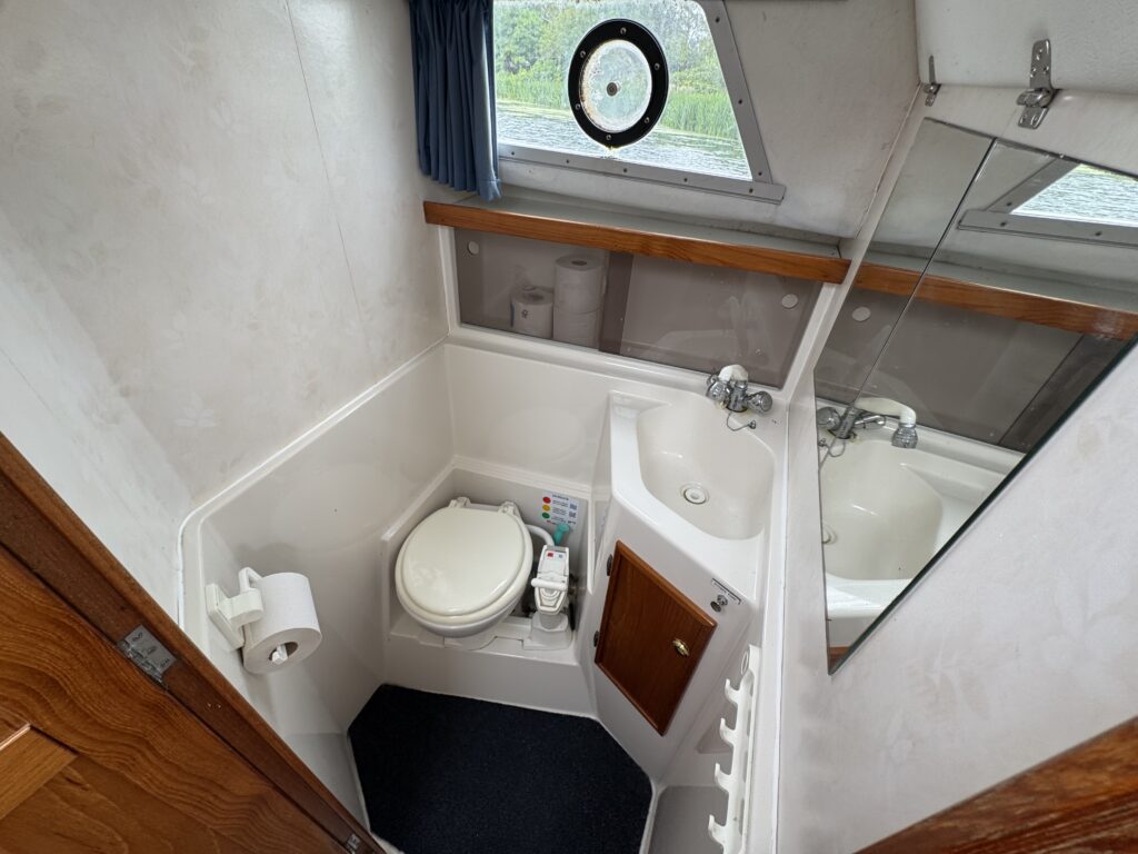 A compact boat bathroom with a toilet, small sink, mirror, and dark floor mat, shown beside a wooden door and a circular window.