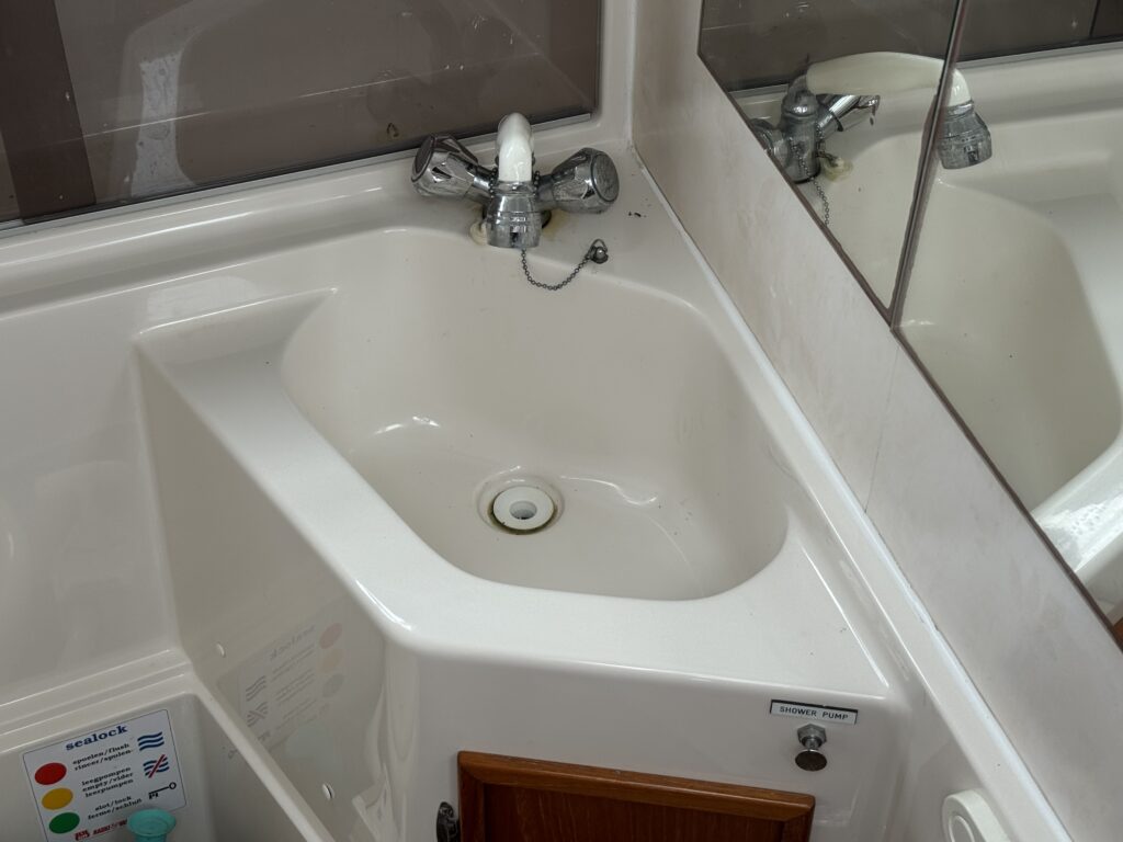 A white boat bathroom sink with chrome faucets and a mirrored wall, shown beside a toilet control panel and a wood cabinet.
