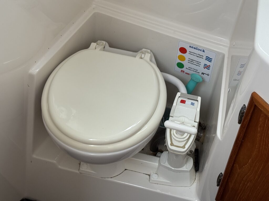 A compact marine toilet installed in a small white cabin compartment, with a manual pump and a Sealoak label beside it.
