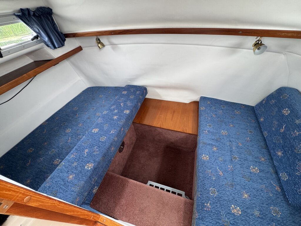 A compact boat cabin with two blue upholstered berths facing each other, a small wooden table between them, and a curtained window along the white curved wall.