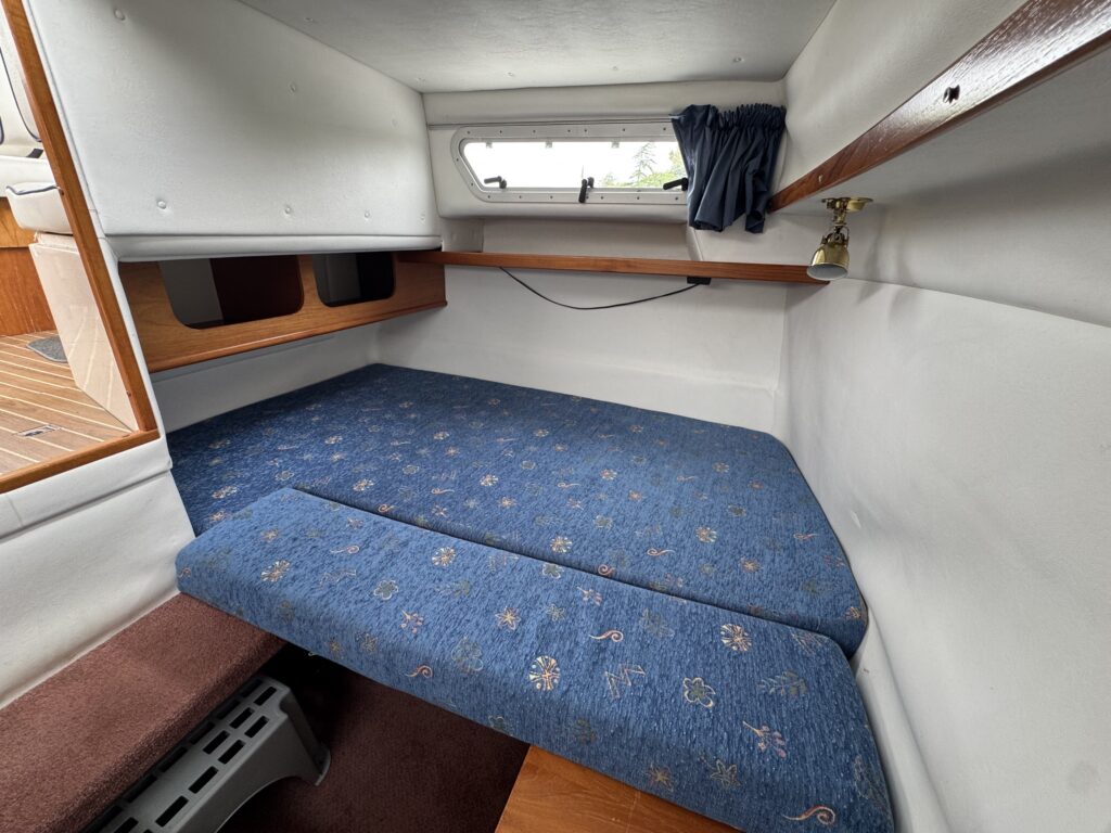 A compact boat cabin with a blue cushioned sleeping berth, white walls, a small window with a dark curtain, and wooden trim.