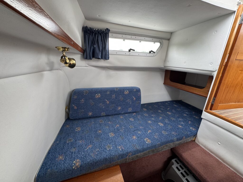A blue upholstered cabin berth with a matching backrest sits inside a small boat interior, with a porthole window, navy curtains, and a wood-trimmed shelf above.