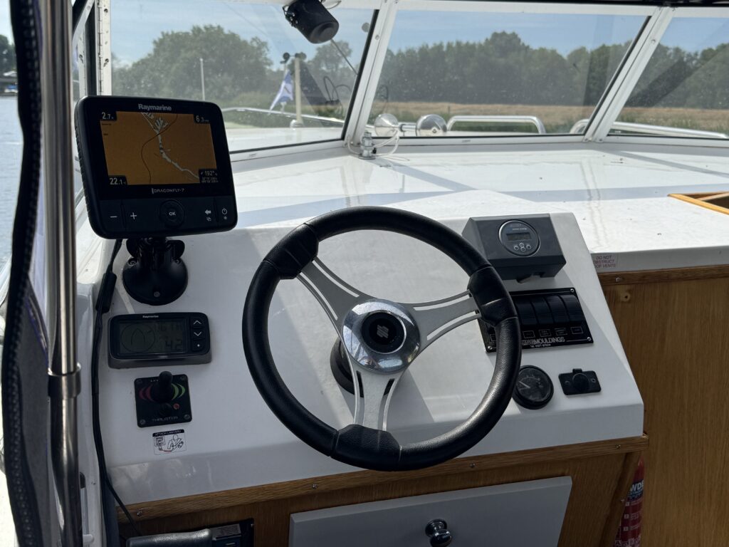 A boat helm with a black steering wheel, GPS display, and control switches, viewed through the windshield with trees in the background.
