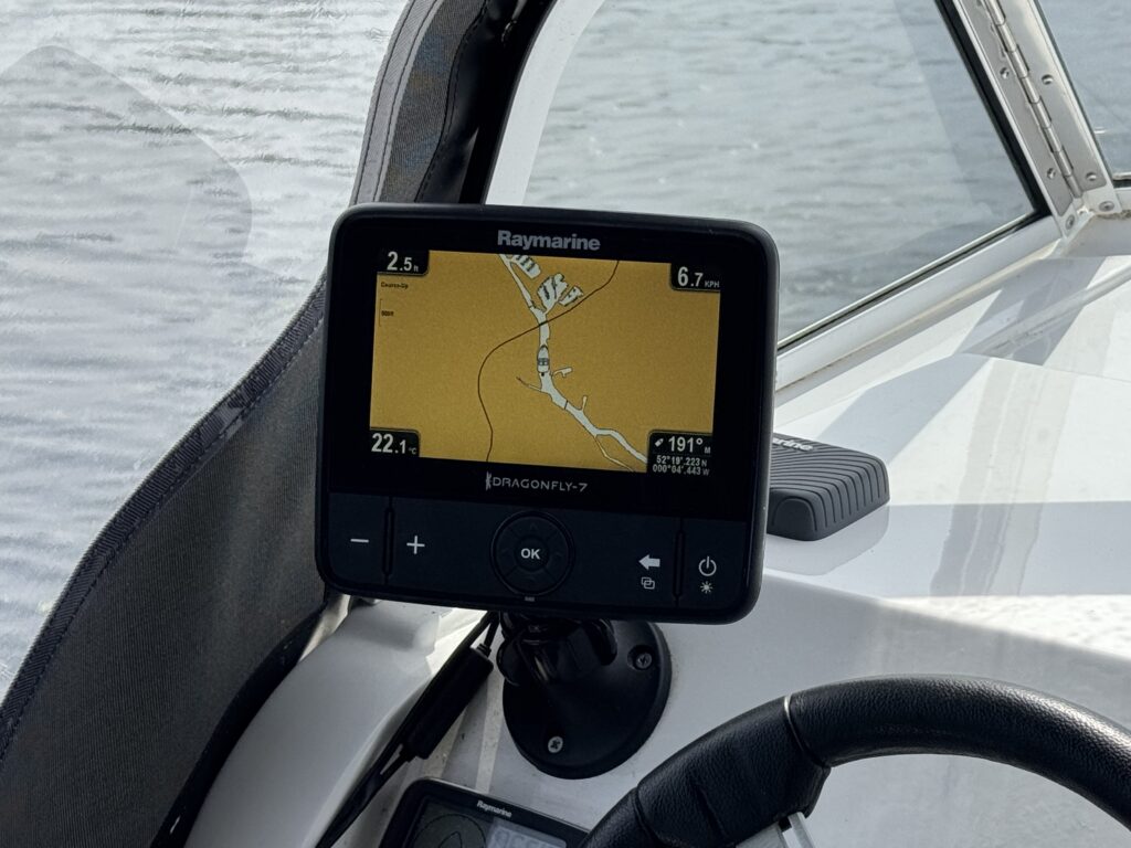 A Raymarine boat navigation display showing a route on a yellow map, mounted beside a steering wheel on a white dashboard.