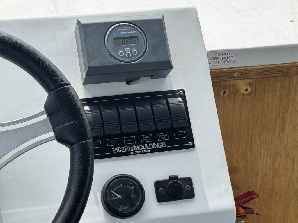 A boat control panel with a steering wheel, digital display, switches, and gauges beside a wood-paneled wall.