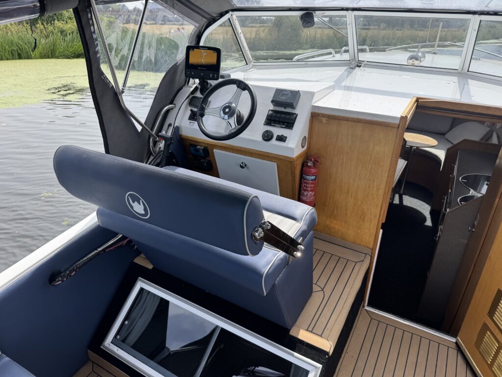 A boat cockpit with a blue helm seat, steering wheel, and navigation display, with water visible beyond the open cabin.