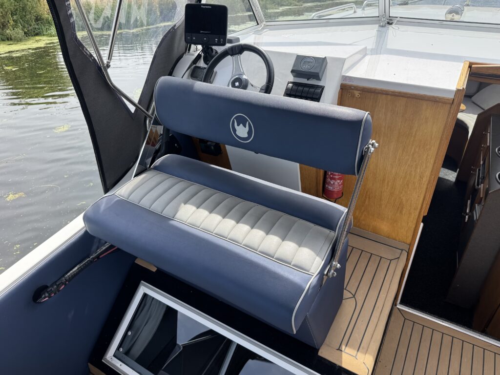 A blue cushioned helm seat on a boat, with a steering wheel, small display, and wooden deck beside the water.