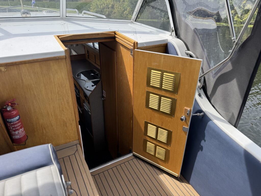 An open wooden cabin door on a boat reveals a narrow interior corridor, with a red fire extinguisher mounted on the left wall and water visible outside.
