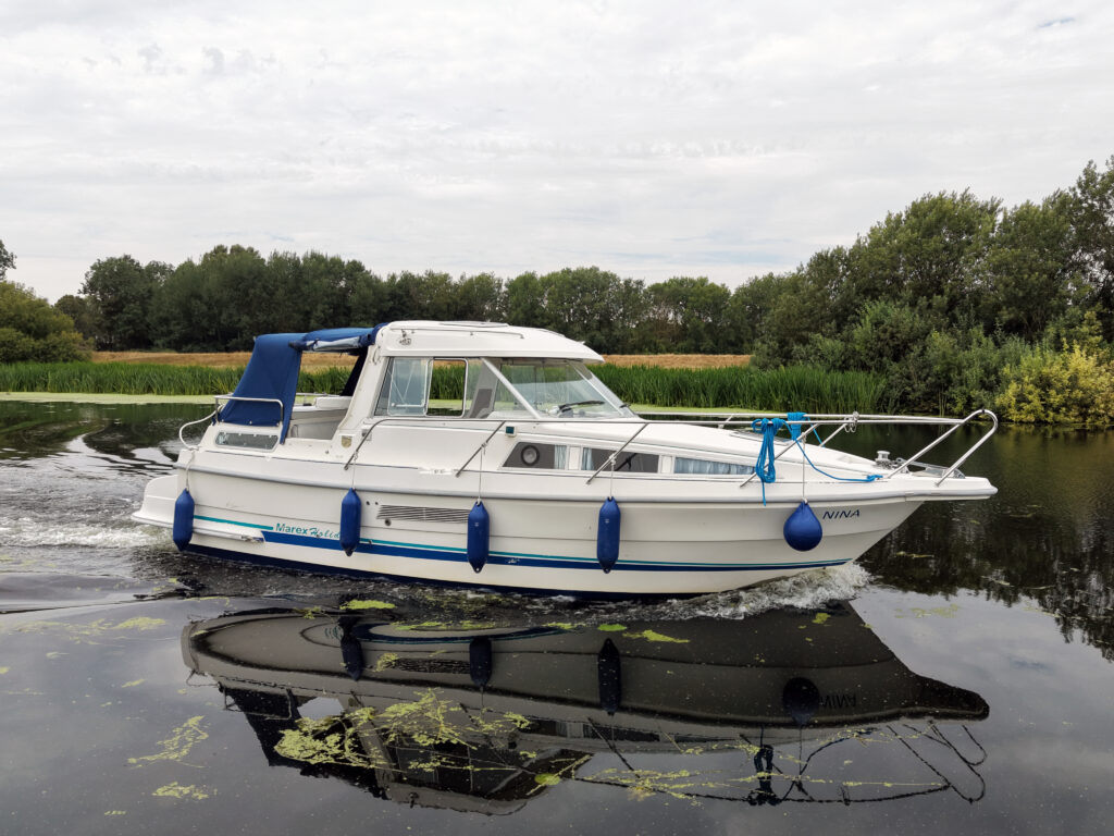 A white Marex 280 Holiday motorboat with blue fenders floats on calm water beside a tree-lined shore, with its reflection visible on the surface.