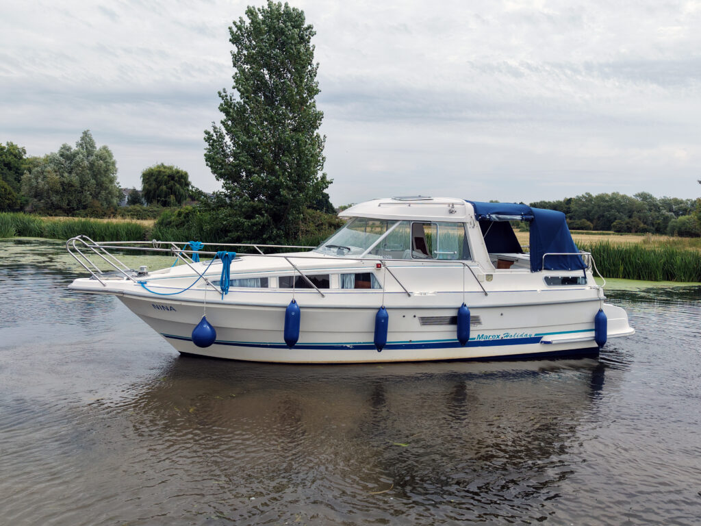 A white Marex 280 Holiday motorboat with blue fenders and a blue canopy floats on calm water beside a grassy riverbank and trees.