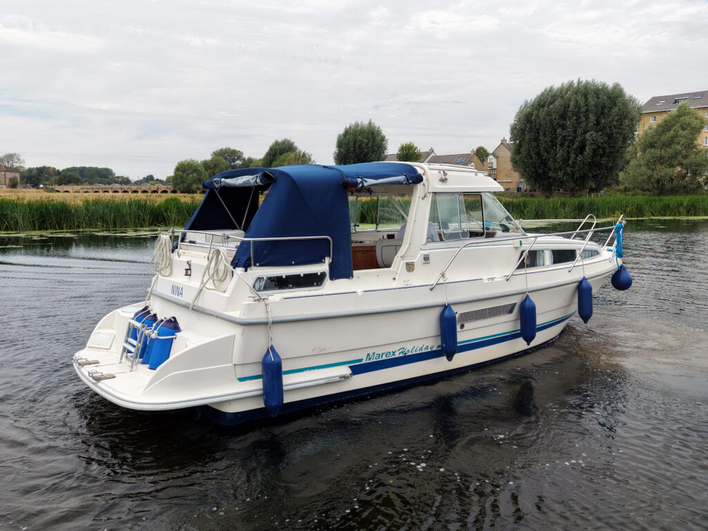 A white Marex 280 Holiday motor yacht with a blue canopy is cruising on a calm river, with reeds, houses, and trees along the bank.