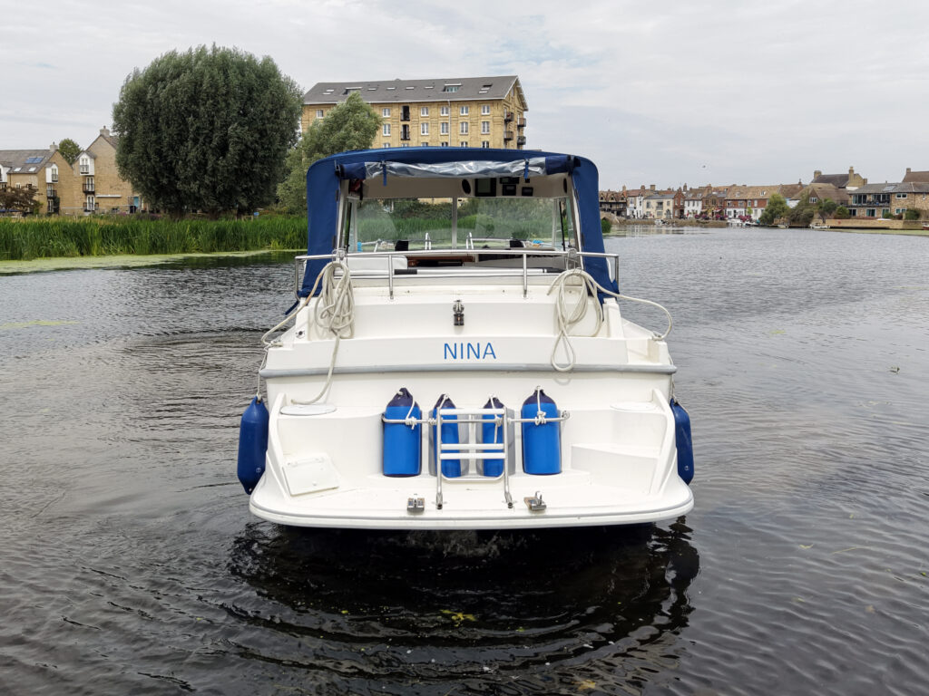 A white Marex 280 Holiday boat named NINA is shown from the stern on calm water, with blue fenders and a blue canopy in front of lakeside buildings.