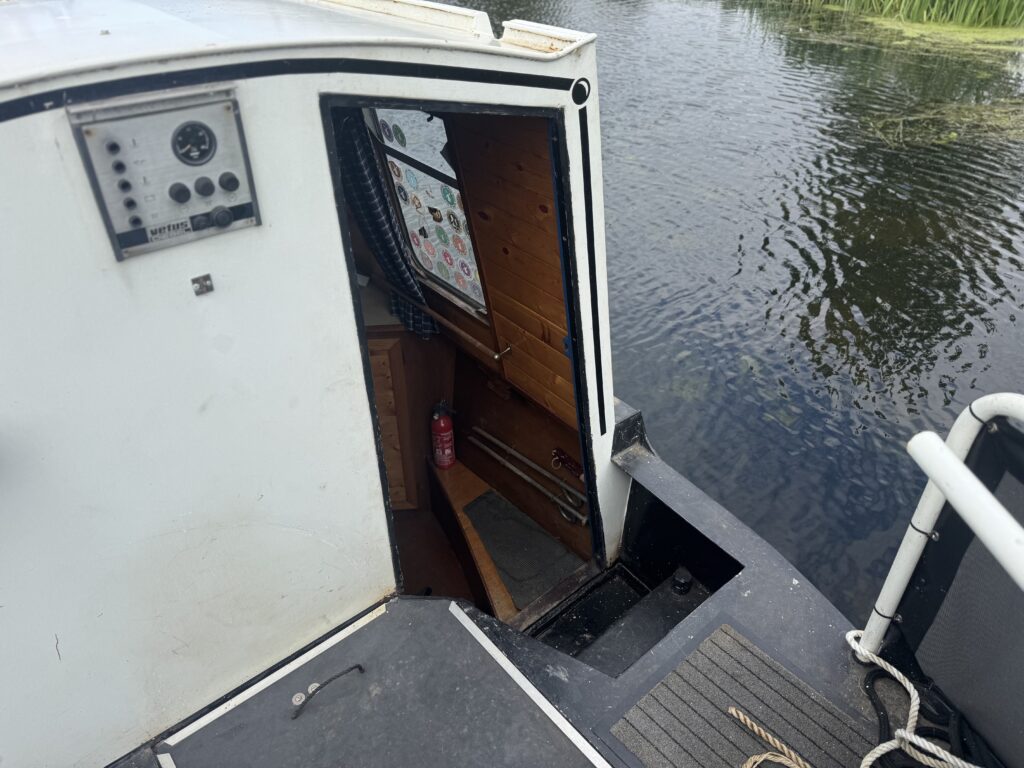 An open narrowboat cabin doorway with a control panel on the left wall and wooden steps inside, beside calm water and a white railing.