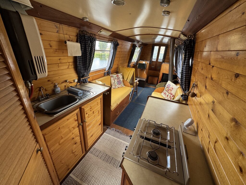 A narrowboat galley with wood-paneled walls, a sink and stove in the foreground, and a seating area with cushions and curtains along the windows.