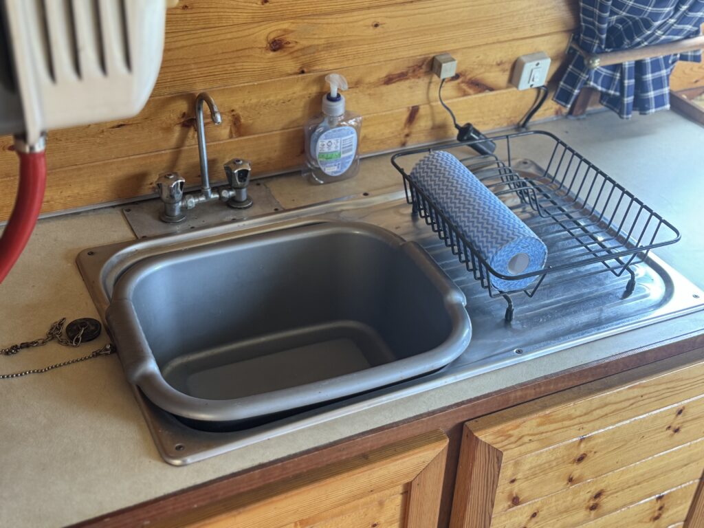 A stainless steel sink with a dish rack and blue paper towel roll sits in a narrowboat galley with wooden cabinets and a wood-paneled wall.