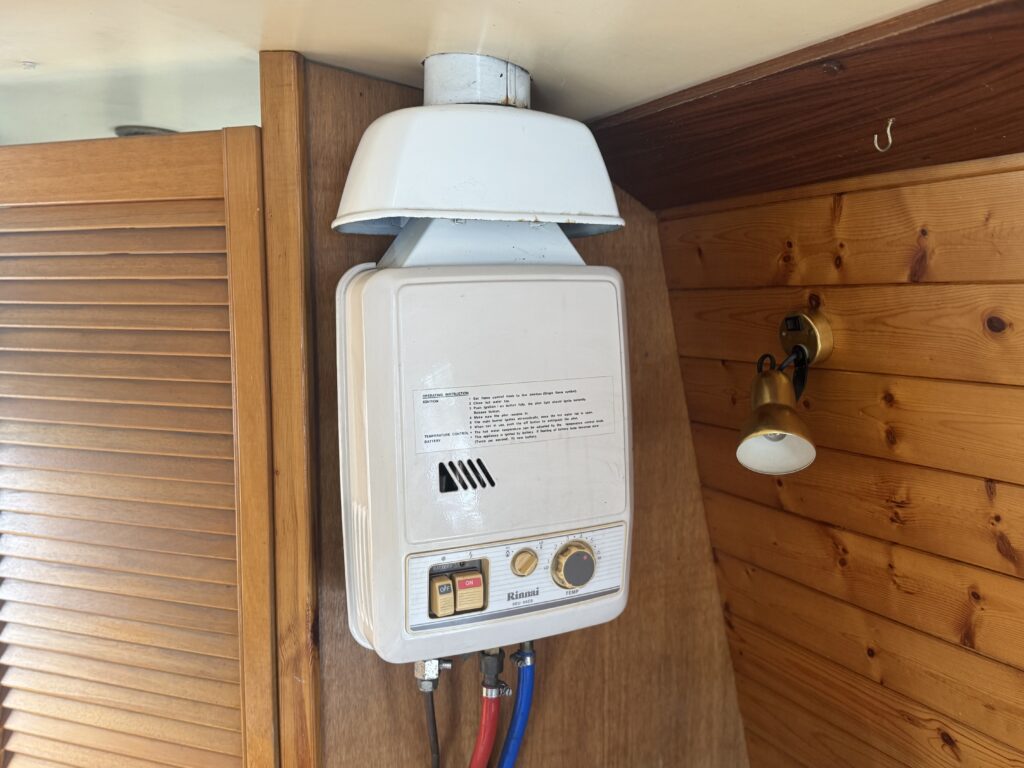 A wall-mounted white gas boiler on a narrowboat, with pipes and control knobs below and wood-paneled walls around it.