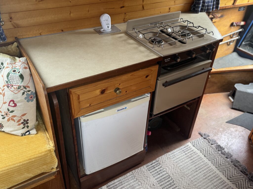 A compact boat kitchenette with a countertop, two-burner stovetop, small oven, and mini fridge, set beside a patterned chair and wood-paneled wall.