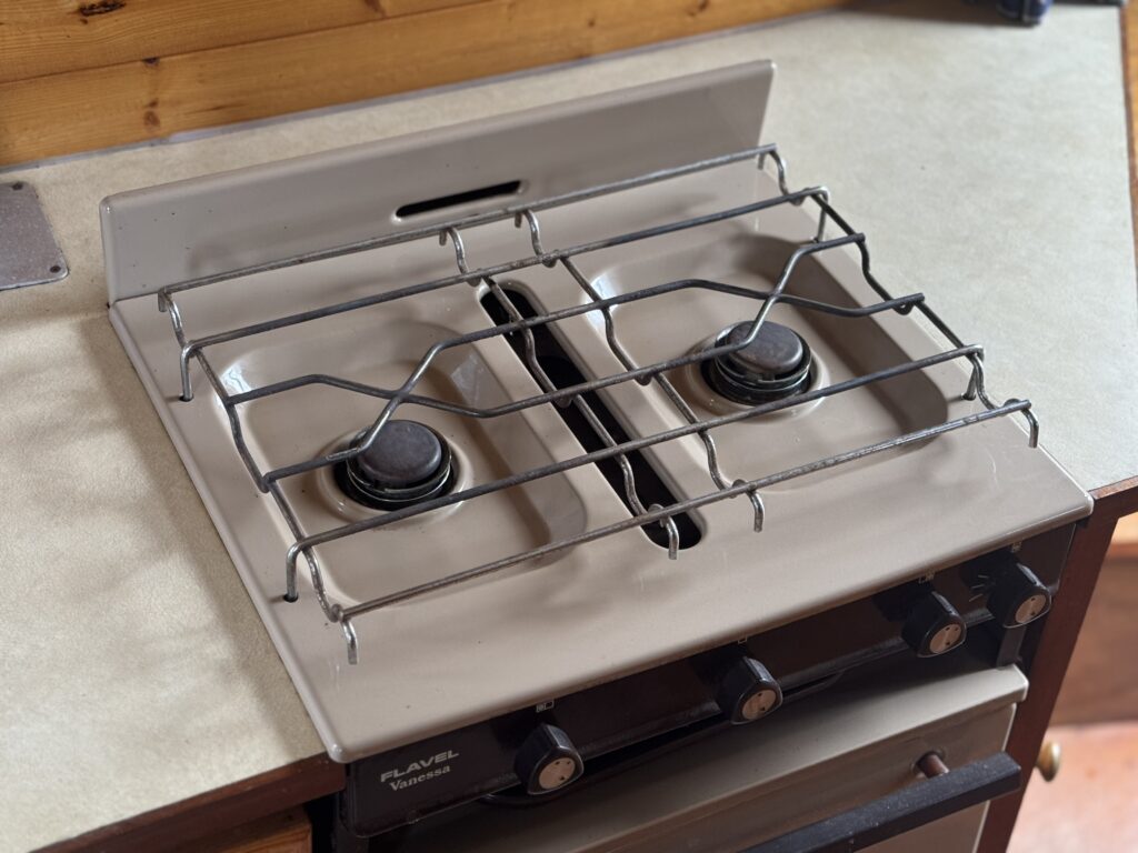 A compact two-burner gas stove with a metal pan support sits on a countertop beside a wooden backsplash.