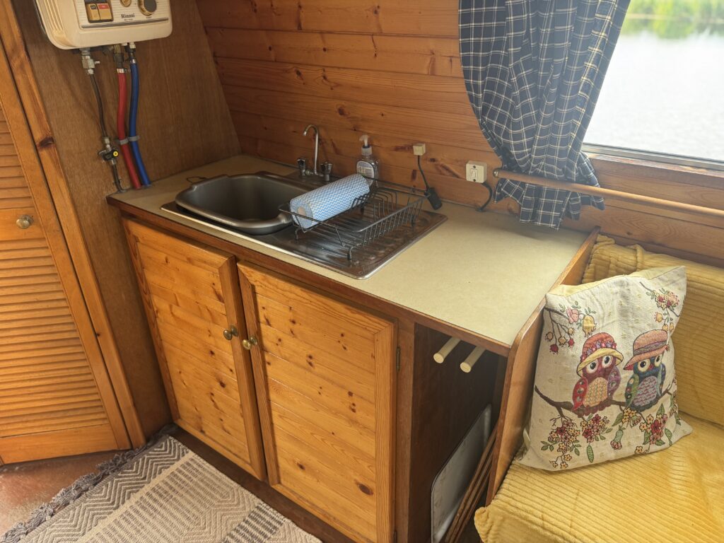 A compact narrowboat kitchen with wooden cabinets, a stainless-steel sink, and a countertop dish rack beside a window with blue curtains.