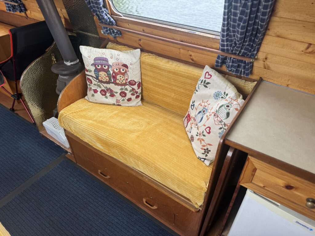 A yellow-upholstered bench seat in a narrowboat cabin with owl-print and floral cushions beside a wood-burning stove and a window with blue curtains.
