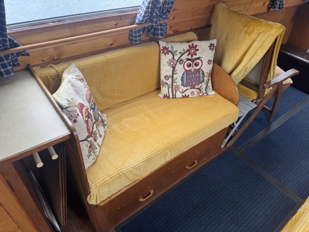 A yellow upholstered bench seat on the Owl Class narrowboat with owl-patterned cushions beside a fold-down wooden table and blue carpet.