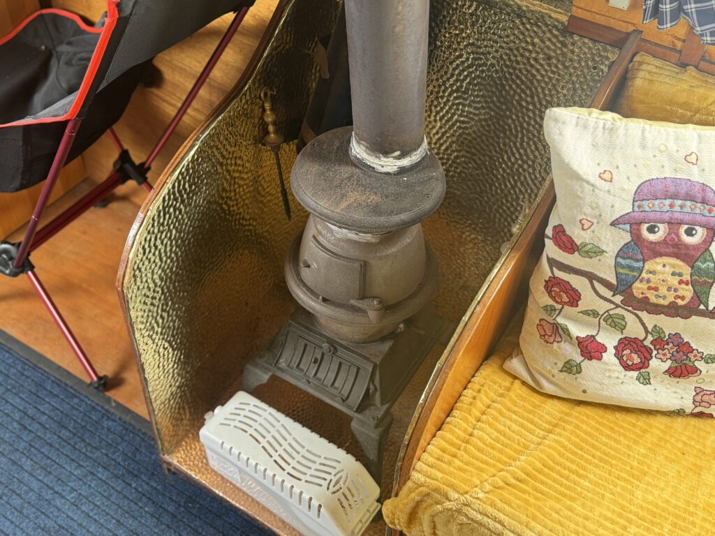 A cast-iron stove with a chimney pipe sits beside a yellow cushion and an owl pillow inside a narrowboat.