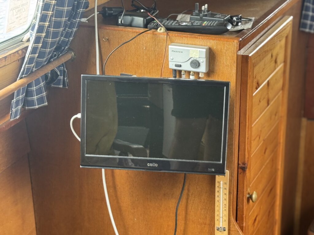 A small flat-screen television mounted on a wooden cabinet in a narrowboat, with cables, a control panel, and a plaid curtain nearby.