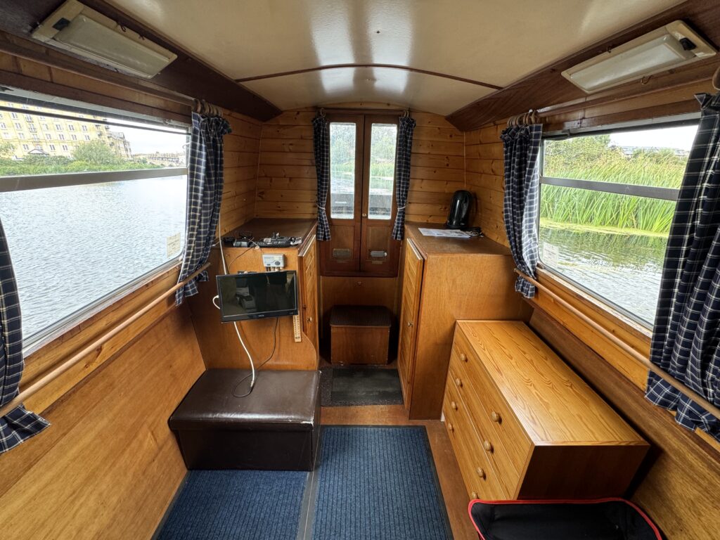 A narrowboat cabin with wood-paneled walls, blue rugs, and bench seating beside large side windows overlooking the water.