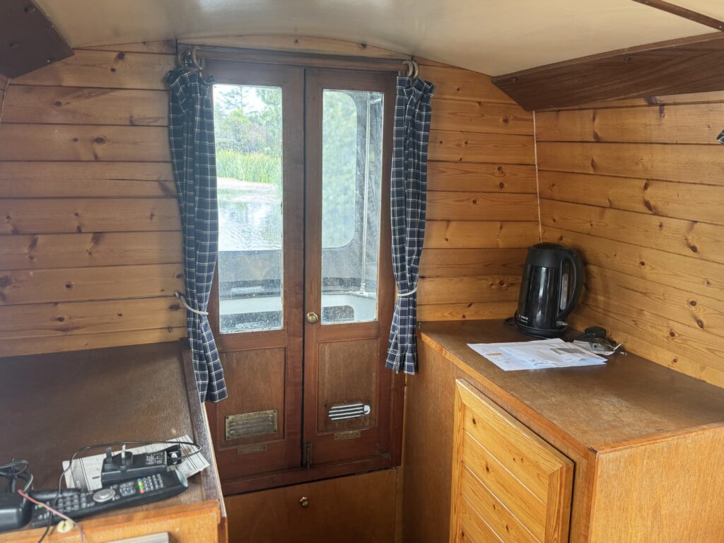 A narrowboat cabin with wooden panel walls, a pair of glass doors with plaid curtains, and a counter holding a black kettle and papers.