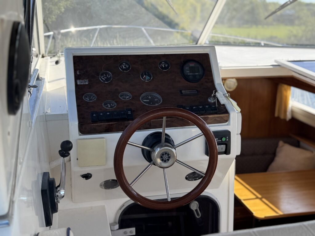 The helm of a Sheerline 950 boat with a wood-rim steering wheel, gauges, and a throttle panel, beside a wooden bench and window with water and shoreline visible outside.