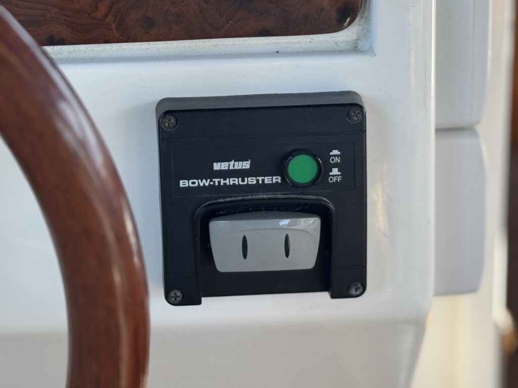 A black Vetus bow-thruster control panel with a green on/off button is mounted on a white surface beside a wooden steering wheel.