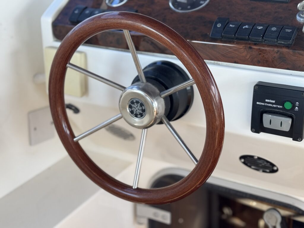 A polished wooden steering wheel with metal spokes is mounted on a cream boat console, with wood trim, switches, and a power outlet in the background.