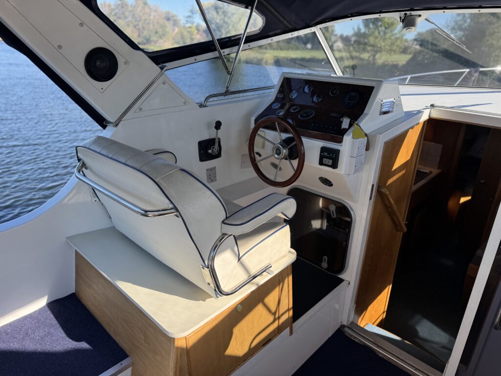 A white boat helm seat with wooden trim and a steering wheel sits beside the dashboard, with water visible through the windshield and an open cabin doorway.