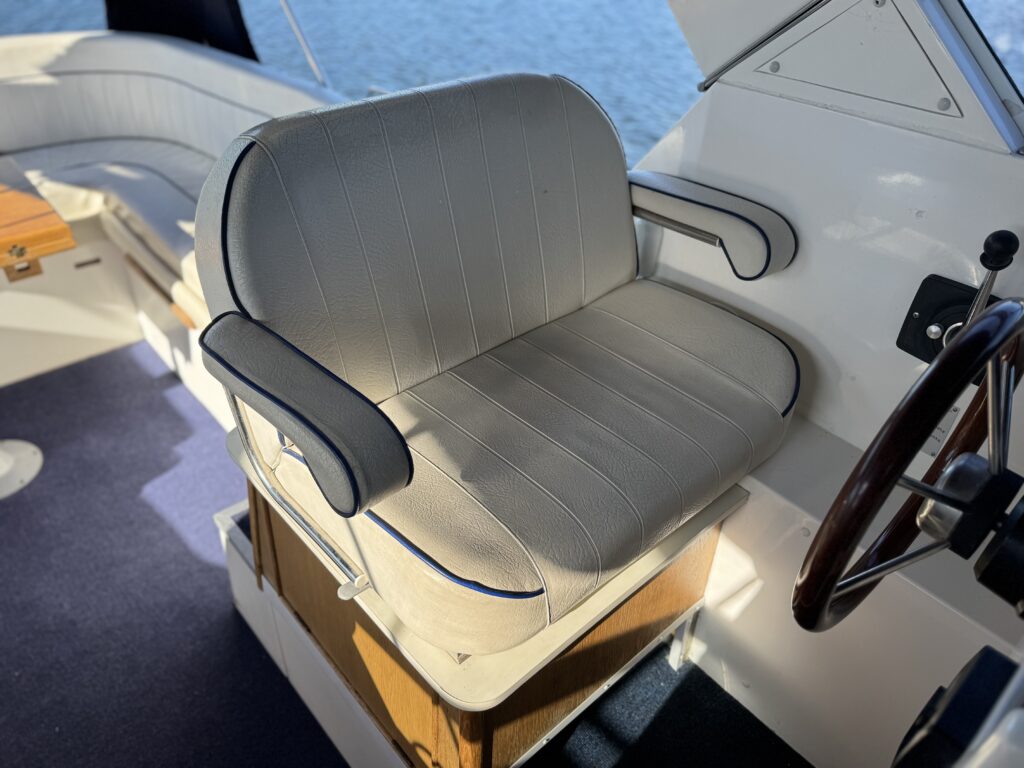 A cushioned gray helm seat with black armrests beside a boat steering wheel, shown on a Sheerline 950 with water visible in the background.
