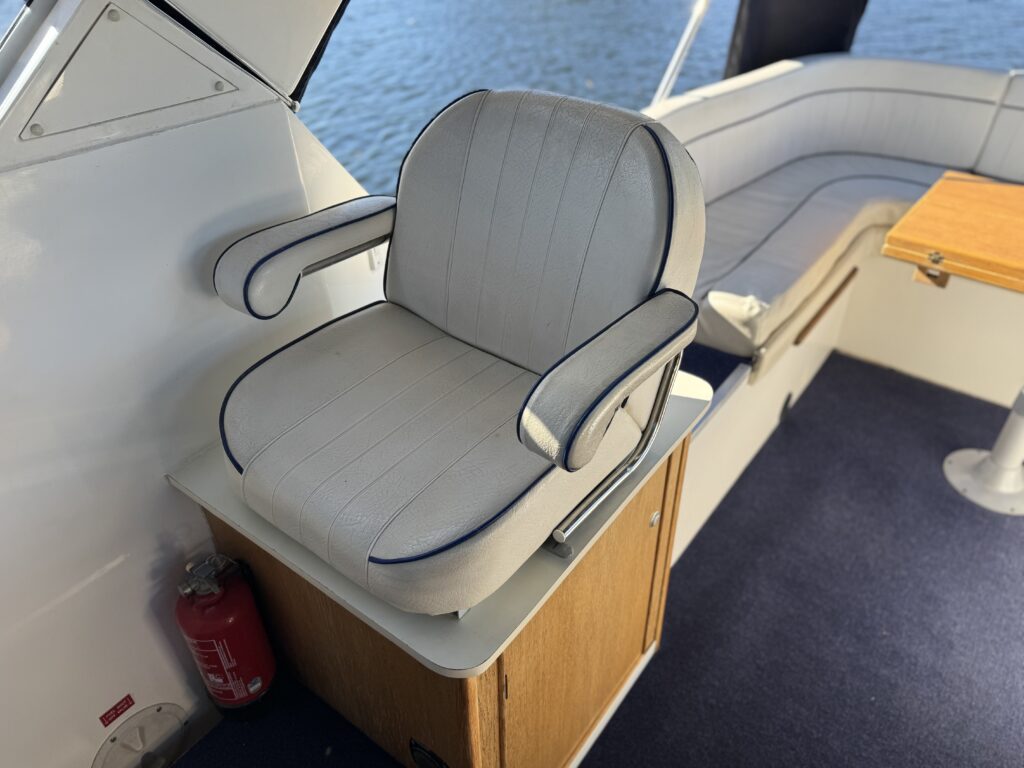 A light gray cushioned helm seat with navy piping sits beside a boat window, with blue carpet, a wooden cabinet, and water visible in the background.