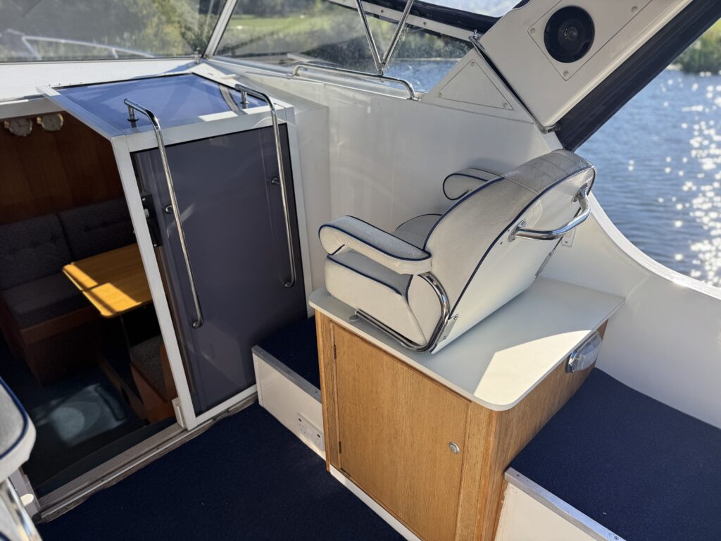 A white cushioned helm seat sits beside a compact cabinet and storage compartment on a boat, with a blue carpeted deck and water visible through the windows.