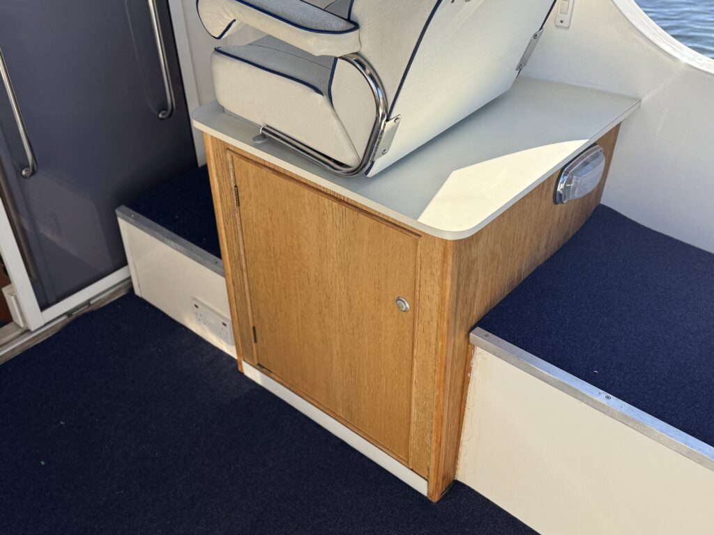 A built-in wooden storage cabinet with a white cushioned seat on top, set between blue upholstered benches on a boat interior.