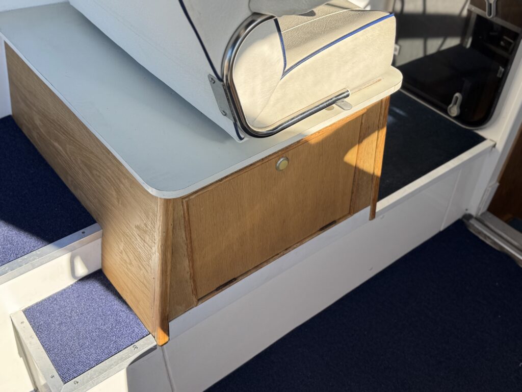 A wooden cabin bench with a cream upholstered cushion and metal railings, set on a boat with blue carpeted flooring.