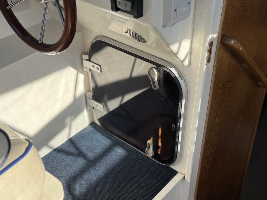 A black-framed access hatch installed in a white cabin wall beside a wooden doorframe, with blue carpeted flooring and part of a steering wheel visible.
