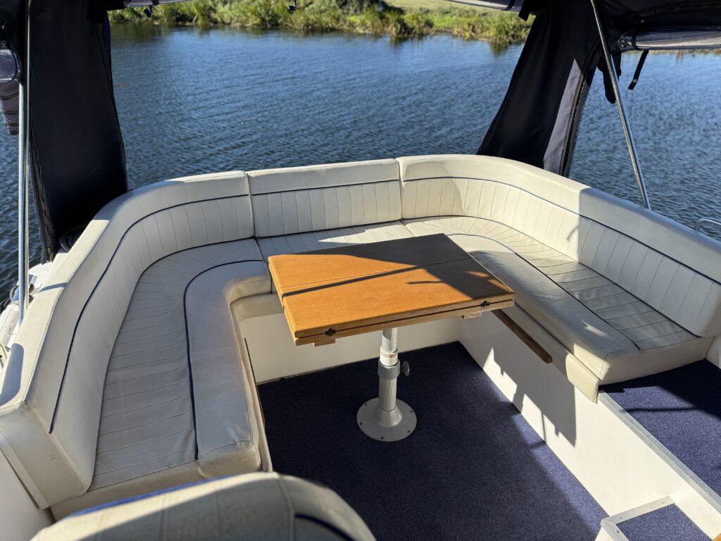 A cream-colored curved bench seating area on a boat with a wooden table in the center, framed by open windows overlooking the water.